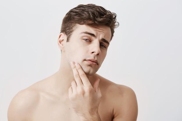 Homem bonito com cabelo escuro, corte de cabelo à moda, olhos azuis, mantém a mão no queixo, parece pensativo, isolado. Cara europeu nu confiante com pele pura e saudável