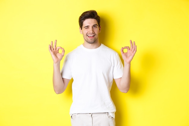 Homem bonito confiante piscando, mostrando sinais de aprovação, como algo bom, em pé sobre um fundo amarelo