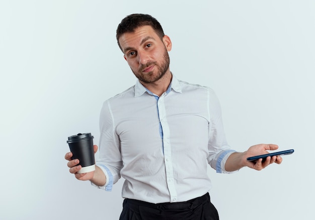 Homem bonito confiante segurando a xícara de café e o telefone isolado na parede branca