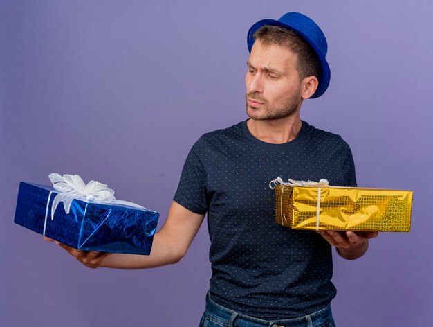 Homem bonito confuso com chapéu azul segura e olha para as caixas de presente isoladas na parede roxa