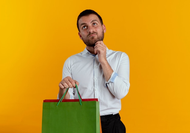 Foto grátis homem bonito confuso segurando sacolas de papel e colocando a mão no queixo, olhando para cima, isolado na parede laranja