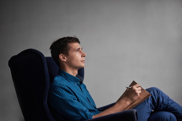 Homem bonito de camisa azul e calça jeans, sentado na cadeira escura e escrevendo ideias no bloco de notas