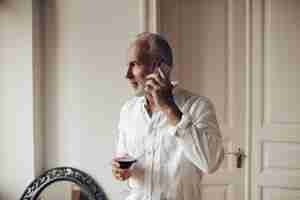 Foto grátis homem bonito de camisa branca fala no telefone e segurando o copo de vinho retrato de cara adulto com barba grisalha e roupa elegante leve com bebida vermelha na mão