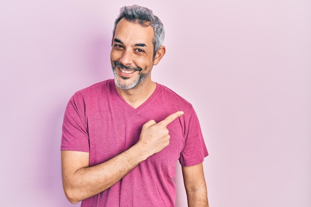 Homem bonito de meia idade com cabelos grisalhos vestindo camiseta rosa casual sorrindo alegre apontando com a mão e o dedo para o lado