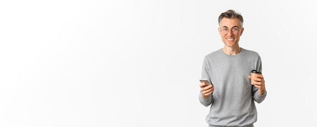 Foto grátis homem bonito de meia-idade com camisola cinzenta e óculos sorrindo feliz bebendo café e usando celular