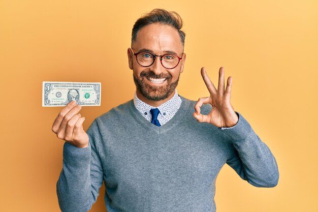 Homem bonito de meia-idade segurando uma nota de 1 dólar fazendo sinal de ok com os dedos sorrindo amigável gesticulando excelente símbolo