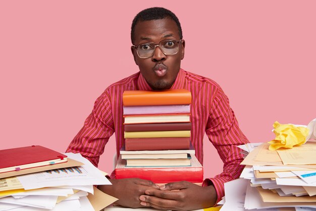 Homem bonito de pele escura faz beicinho, segura uma pilha de livros, bagunça a mesa, usa óculos e camisa listrada