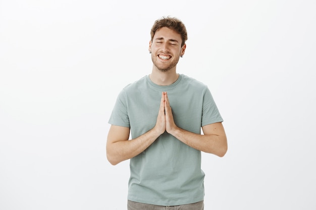 Foto grátis homem bonito de saída positivo em uma camiseta casual, segurando as mãos em uma pirra e sorrindo alegremente, fechando os olhos e sonhando, sendo uma pessoa religiosa e falando com deus