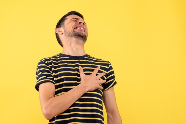 Homem bonito de vista frontal em uma camiseta listrada em preto e branco segurando o peito com dor em um fundo amarelo isolado