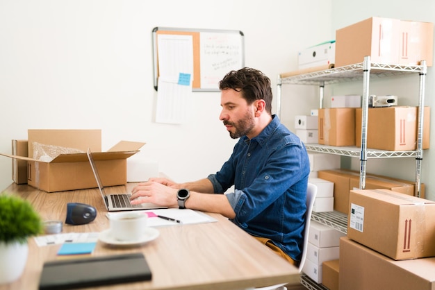 Homem bonito digitando em um laptop e postando em sua loja online os novos produtos de sua inicialização de negócios