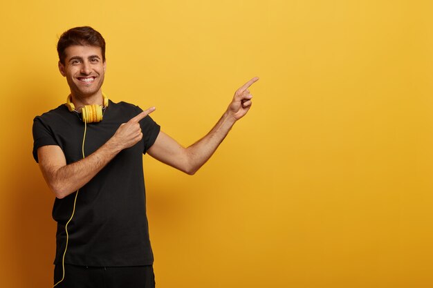 Homem bonito e alegre aponta para o espaço da cópia, vestido com roupas pretas, usa fone de ouvido, sorri com os dentes, demonstra propaganda, isolado sobre fundo amarelo