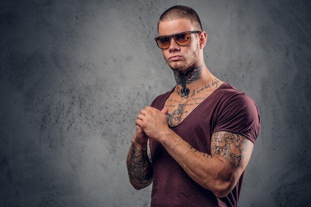 Homem bonito e atlético em óculos de sol com tatuagens nos braços e pescoço posando sobre fundo artístico cinza em um estúdio.