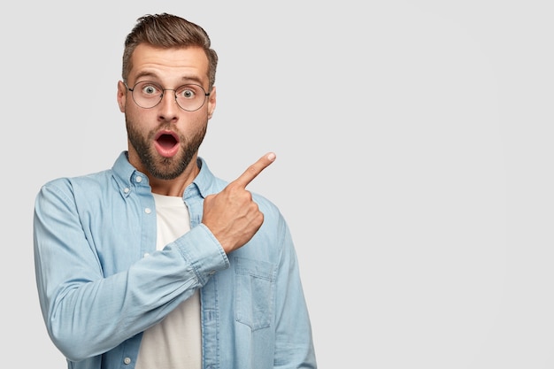 Foto grátis homem bonito e barbado, atordoado, com expressão facial de choque, vestido com uma camisa da moda, aponta para o canto superior direito