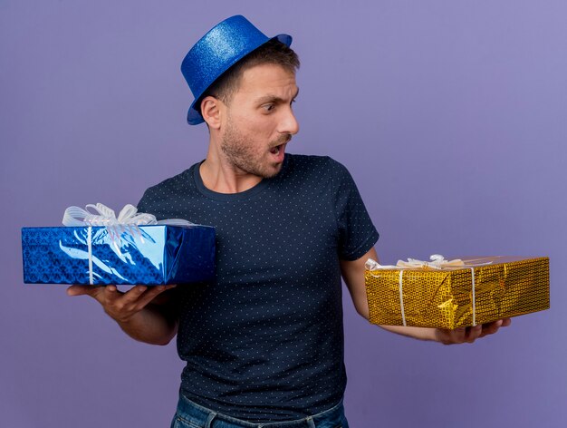 Homem bonito e chocado com chapéu azul segurando e olhando para caixas de presente isoladas em um fundo roxo com espaço de cópia