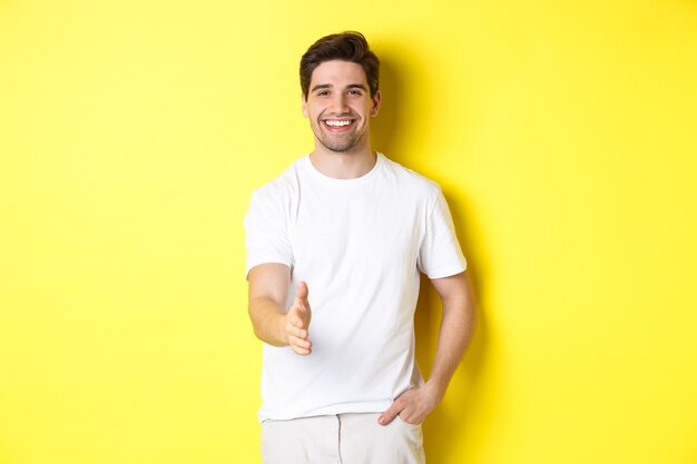 Homem bonito e confiante, estendendo a mão para um aperto de mão, cumprimentando-o, dizendo olá, em pé na camiseta branca sobre fundo amarelo.