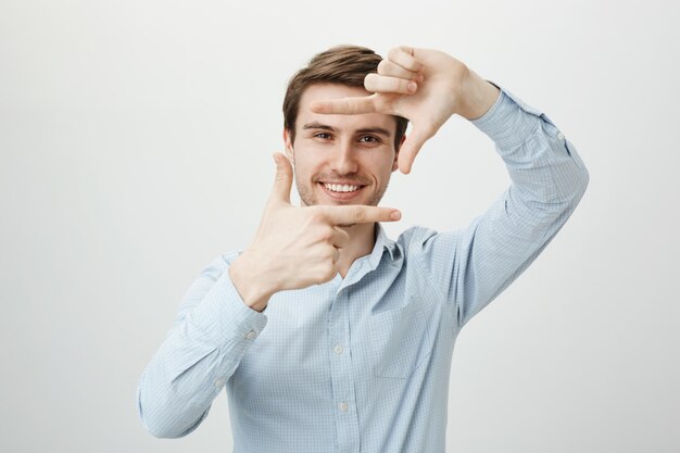 Homem bonito e criativo sorrindo, mostrando molduras de mão, momento de captura