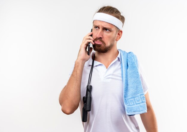 Homem bonito e esportivo confuso usando bandana e pulseiras, falando no telefone com uma toalha e pular corda nos ombros, olhando para o lado isolado na parede branca com espaço de cópia