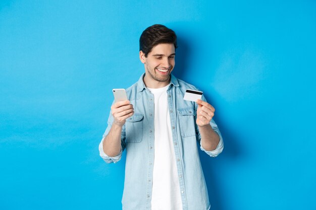 Homem bonito e feliz pagando algo online, segurando o cartão de crédito e o celular, compra na internet, em pé sobre um fundo azul.