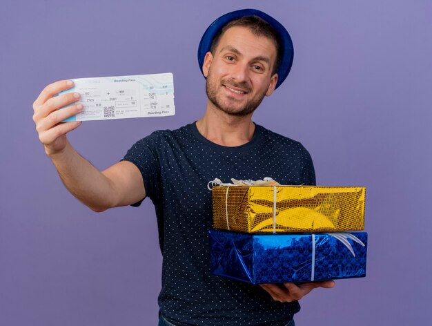 Homem bonito e sorridente caucasiano com chapéu azul segurando caixas de presente e passagem aérea isoladas em um fundo roxo com espaço de cópia