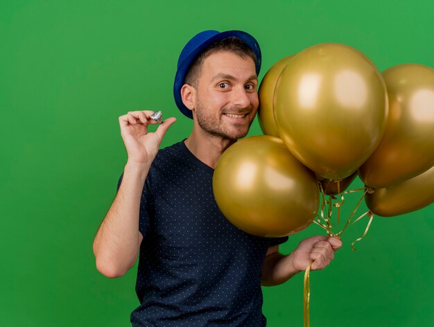 Homem bonito e sorridente caucasiano com chapéu de festa azul segurando balões de hélio e apito de festa isolado em um fundo verde com espaço de cópia