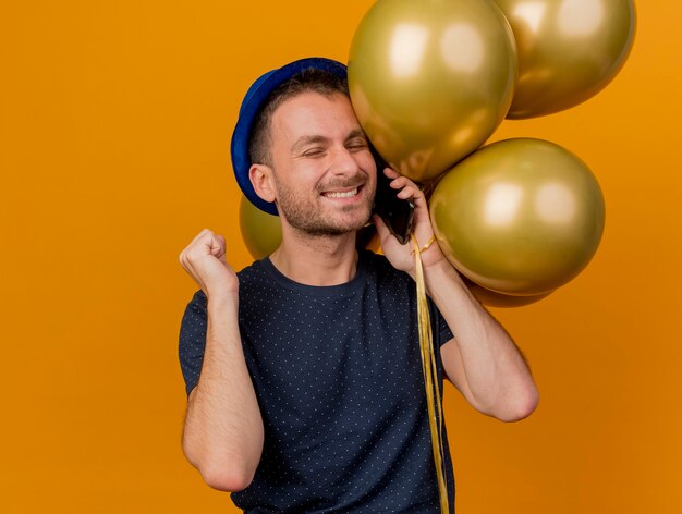 Homem bonito e sorridente, caucasiano, usando chapéu de festa azul, mantém o punho fechado e balões de hélio falando no telefone, isolado em um fundo laranja com espaço de cópia