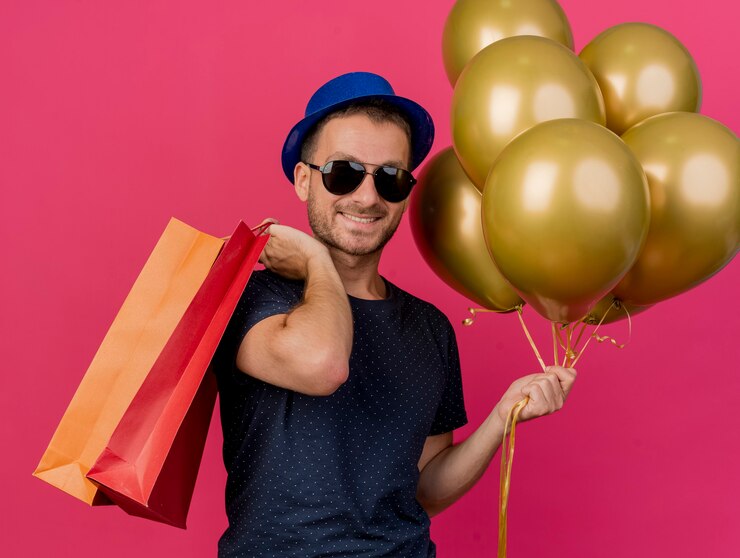 Foto grátis homem bonito e sorridente, caucasiano, usando óculos de sol e chapéu de festa azul, segurando balões de hélio e sacolas de papel isoladas em um fundo rosa com espaço de cópia