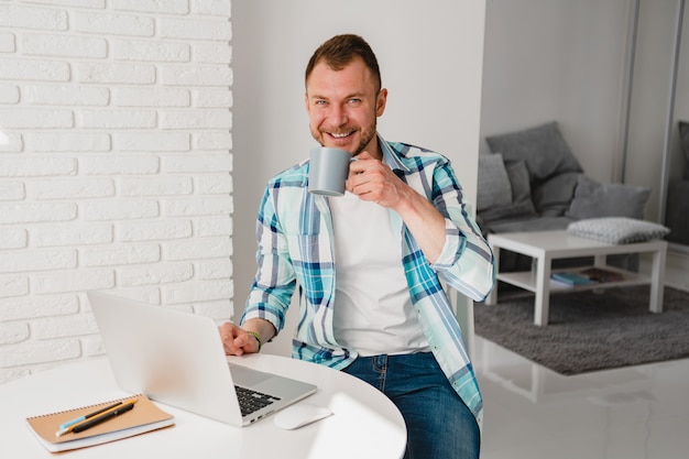 Homem bonito e sorridente com uma camisa, sentado na cozinha em casa, à mesa, trabalhando online no laptop de casa