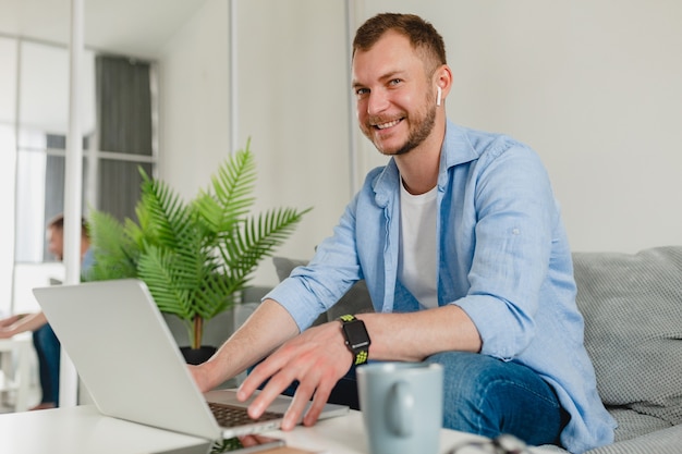 Homem bonito e sorridente com uma camisa sentado relaxado no sofá em casa à mesa trabalhando online no laptop de casa