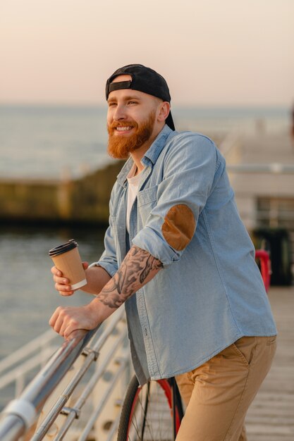 Homem bonito e sorridente hipster barbudo ruivo vestindo camisa jeans e boné com bicicleta no nascer do sol da manhã à beira-mar bebendo café, viajante saudável estilo de vida ativo