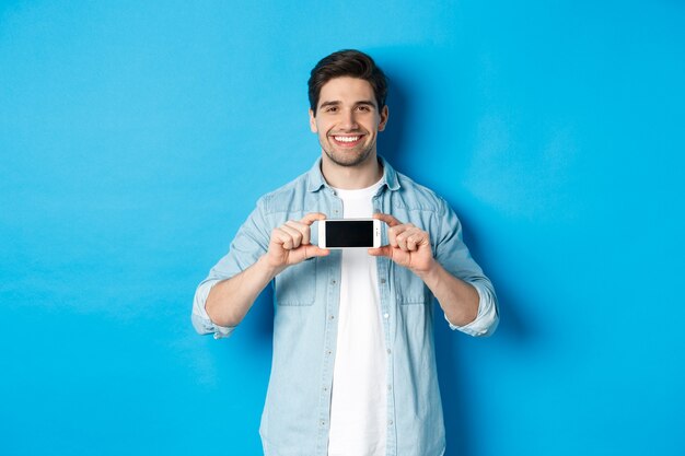 Homem bonito e sorridente mostrando a tela do smartphone, em pé contra um fundo azul para espaço de cópia.