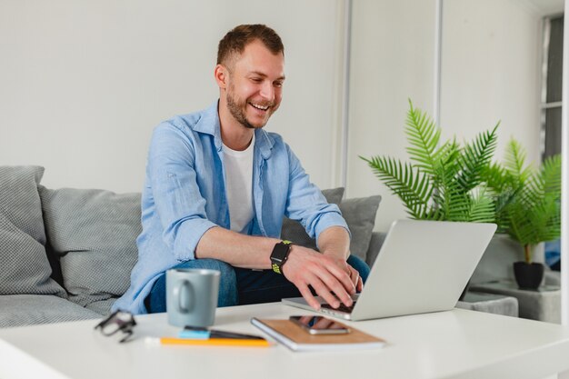 Homem bonito e sorridente sentado no sofá, bebendo chá em casa, à mesa, trabalhando online no laptop de casa