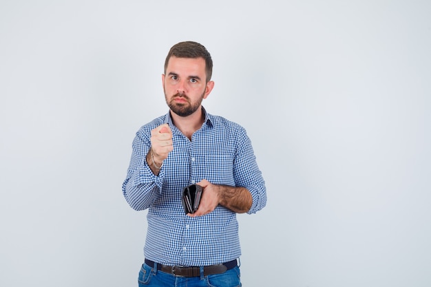 Homem bonito em camisa, jeans segurando a carteira, mostrando o gesto de figo e olhando sério, vista frontal.