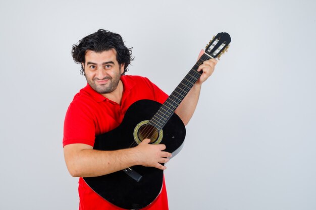 Homem bonito em t-shirt vermelha tocando guitarra e parecendo feliz, vista frontal.