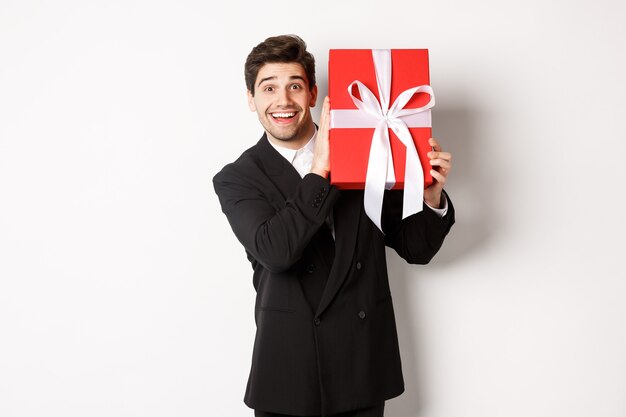 Homem bonito em terno preto, recebendo o presente de Natal, sorrindo espantado, em pé contra um fundo branco.