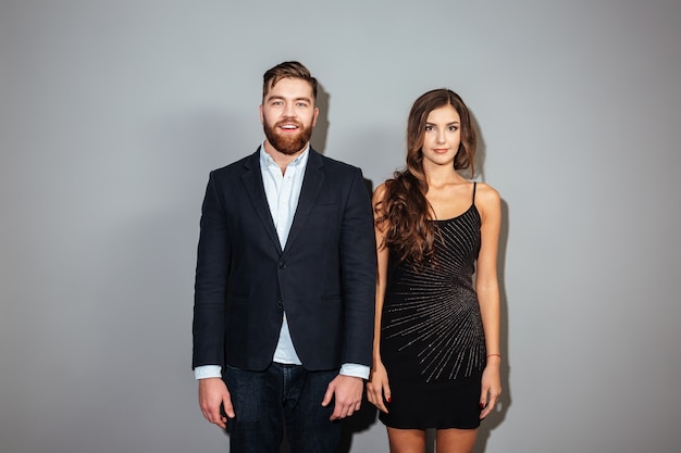 Homem bonito em um terno elegante e uma mulher atraente em um vestido preto em pé e olhando para a frente sobre a parede cinza Foto Premium