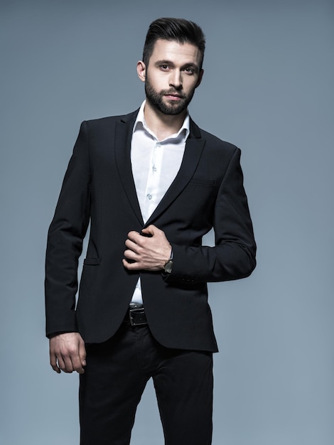 Homem bonito em um terno preto com camisa branca - posando cara atraente com penteado da moda. Homem confiante com barba curta. Menino adulto com cabelo castanho. Retrato completo.