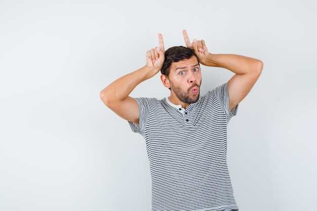 Homem bonito em uma camiseta, mantendo os dedos como chifres de touro, fazendo beicinho nos lábios e olhando bem-humorado, vista frontal.