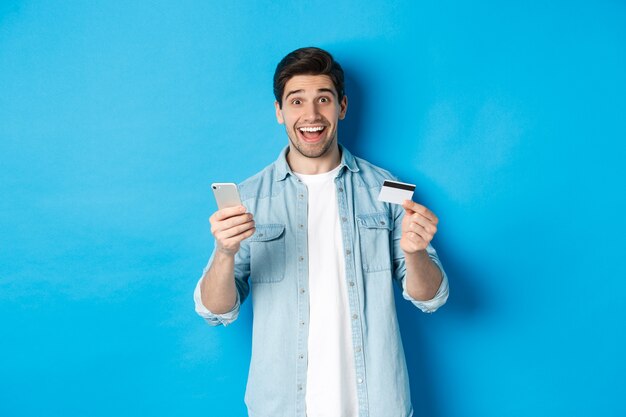 Homem bonito espantado fazendo compras online, segurando o celular e o cartão de crédito, sorrindo enquanto paga a compra pela internet, em pé sobre um fundo azul.