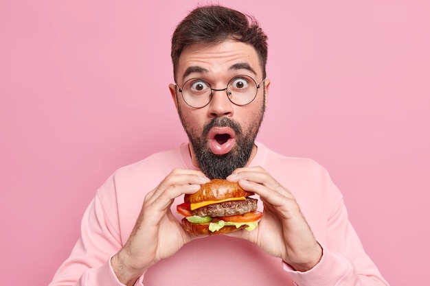 Homem bonito estupefato posa com comida deliciosa e apetitosa de fast food segura hambúrguer saboroso e usa óculos redondos.