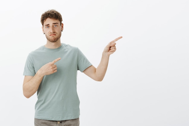 Foto grátis homem bonito indiferente e não impressionado com cerdas nos brincos, apontando para a direita com o dedo indicador e sorrindo