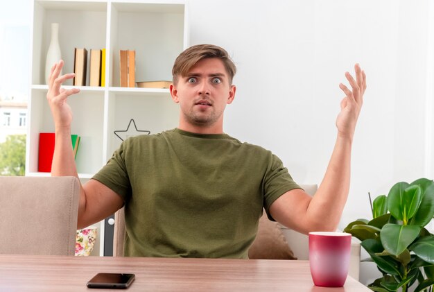 Homem bonito jovem loiro chocado sentado à mesa com o telefone e a xícara, levantando as mãos e olhando para a câmera dentro da sala de estar