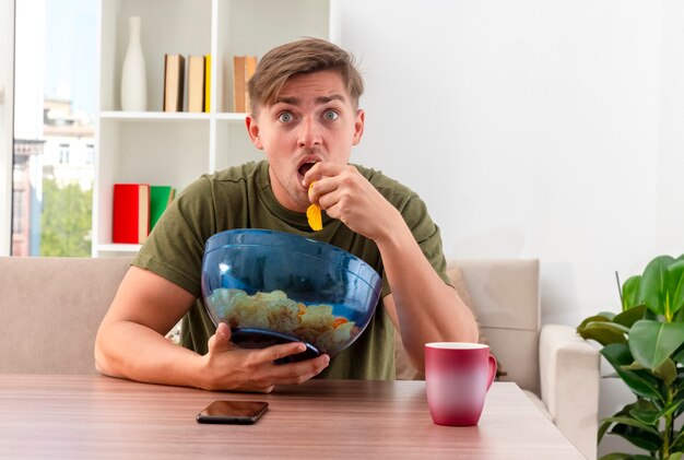 Homem bonito jovem loiro chocado sentado à mesa com uma xícara e um telefone segurando uma tigela de batatas fritas e comendo dentro da sala de estar