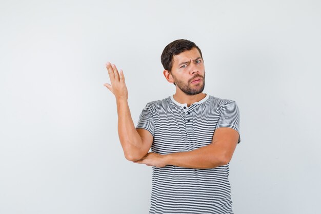 Homem bonito, levantando a mão de forma questionadora em t-shirt e parecendo perplexo, vista frontal.