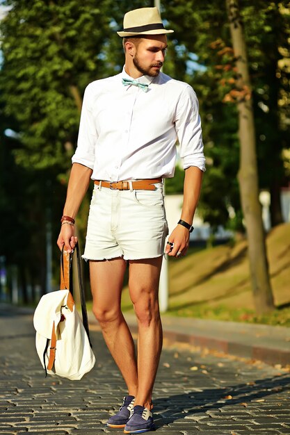 Homem bonito modelo hipster em roupas de verão elegante posando de chapéu com bolsa