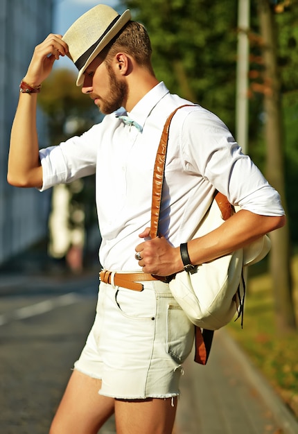 Homem bonito modelo hipster em roupas de verão elegante posando de chapéu com bolsa