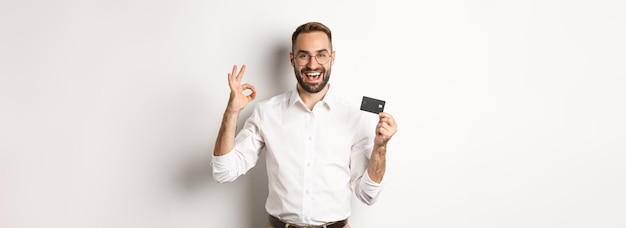 Homem bonito mostrando seu cartão de crédito e sinal de ok recomendando banco em pé sobre fundo branco