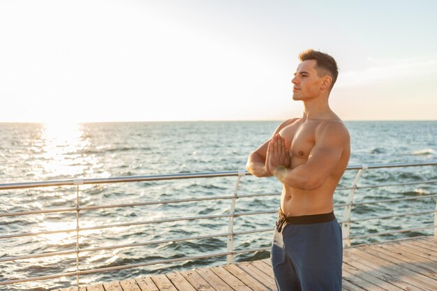 Homem bonito musculoso com torso nu medita na praia ao nascer do sol