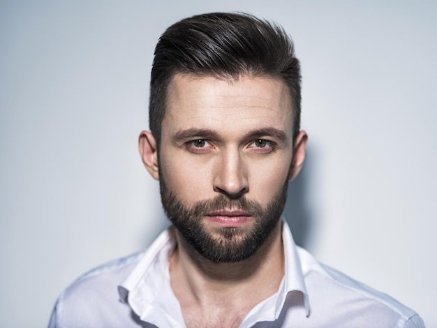 Homem bonito na camisa branca, posando. Cara atraente com penteado da moda. Homem confiante com barba curta. Menino adulto com cabelo castanho. Retrato do close up.