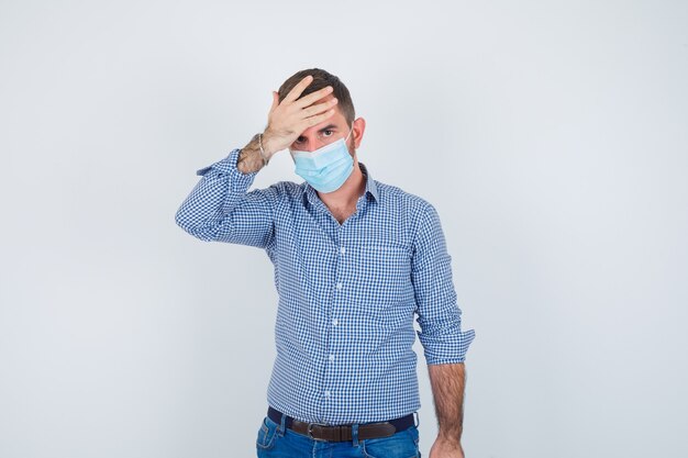 Homem bonito na camisa, jeans, máscara, segurando a mão na cabeça, tendo dor de cabeça e parecendo exausto, vista frontal.