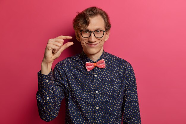 Homem bonito nerd positivo faz pequenos sinais com os dedos, mede algo minúsculo, explica o quanto precisa de algo, vestido formalmente, isolado na parede rosa. Muito pouco